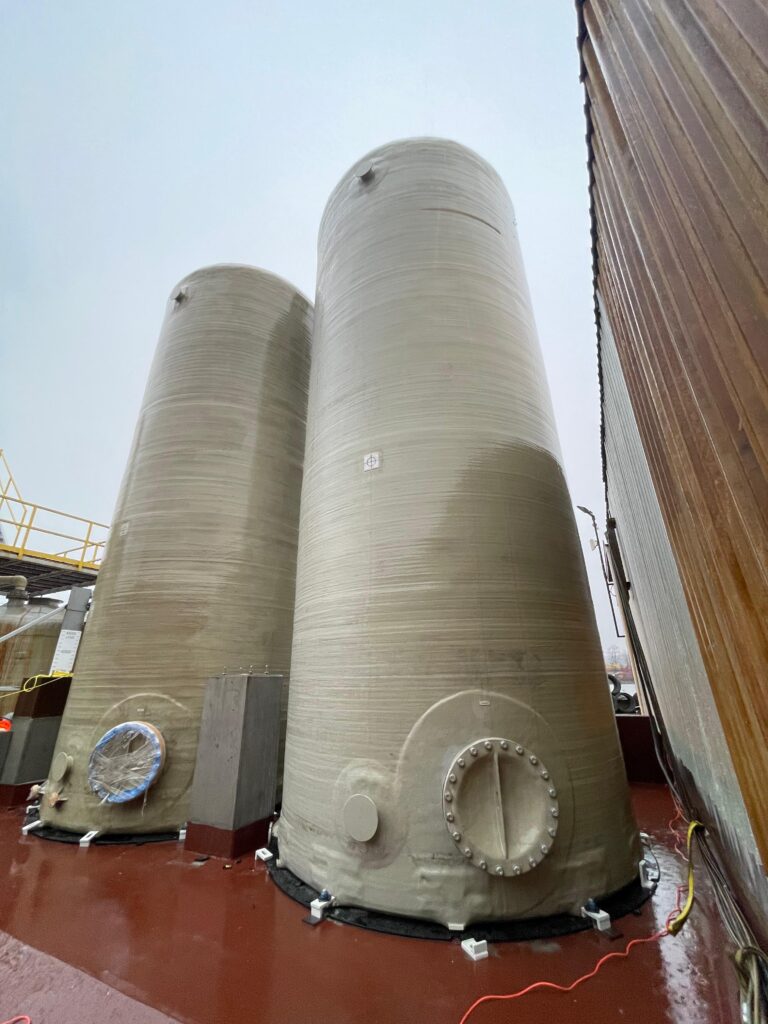 Close-up of two large FRP industrial tanks on a platform, showing circular manway openings and base connection details.